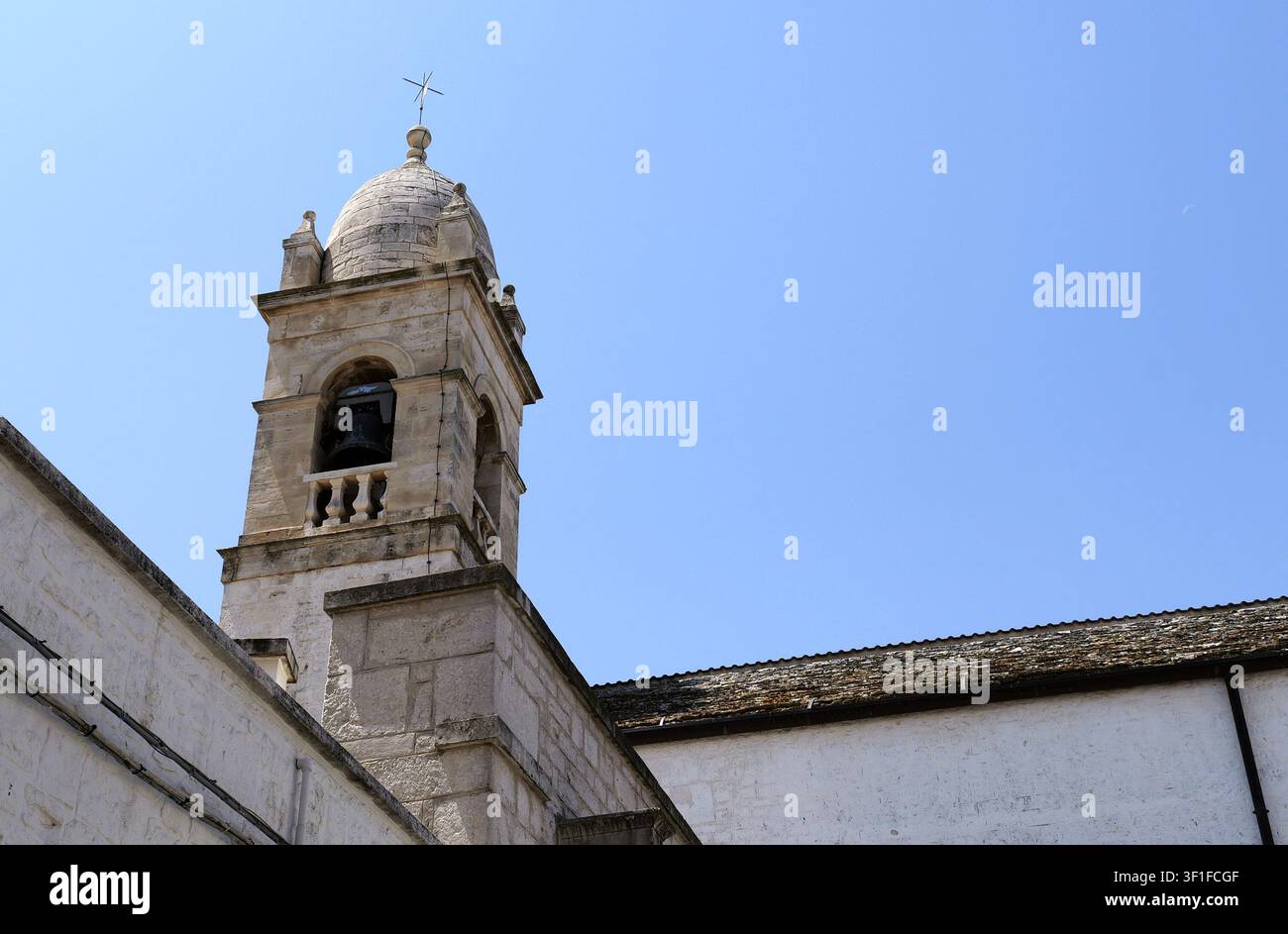 Campanile in pietra con una campana e una croce, parte di una struttura chiesa storica sotto un cielo estivo sereno. Italia, Puglia, Alberobello, 19 luglio 2025 Foto Stock