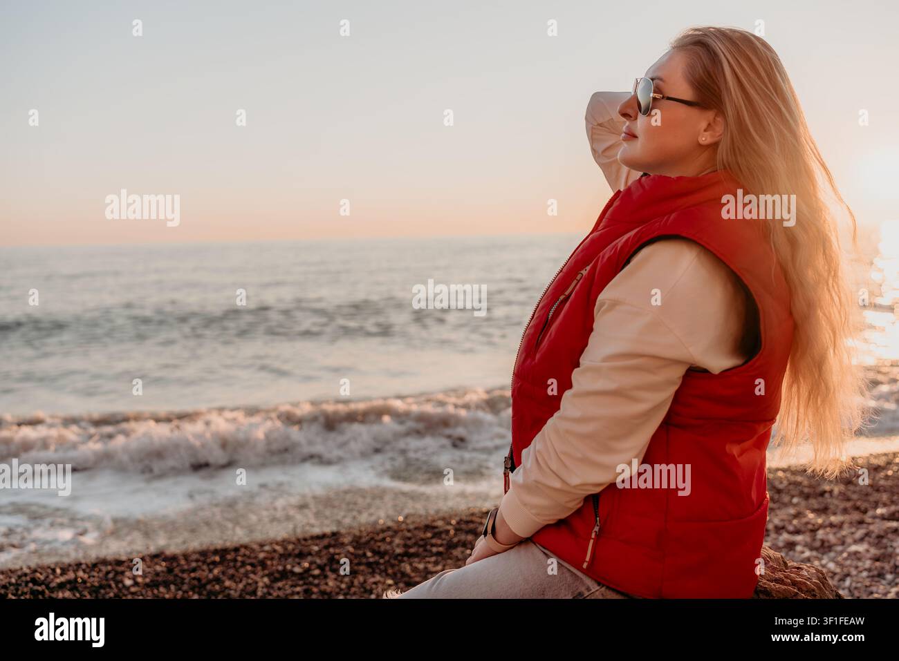 Donna al tramonto sulla spiaggia seduta sulla riva di ciottoli in un gilet rosso che gode di una vista pacifica sull'oceano con spazio per il testo Foto Stock