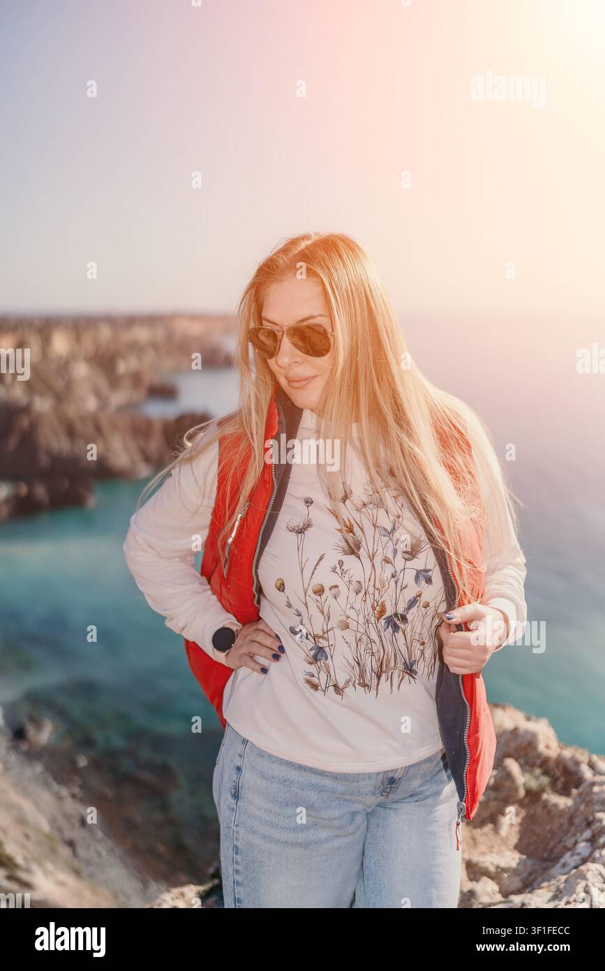 Donna in viaggio lungo la costa in piedi su una scogliera rocciosa che si affaccia sul mare blu al tramonto, indossa abiti casual da esterno e occhiali da sole Foto Stock
