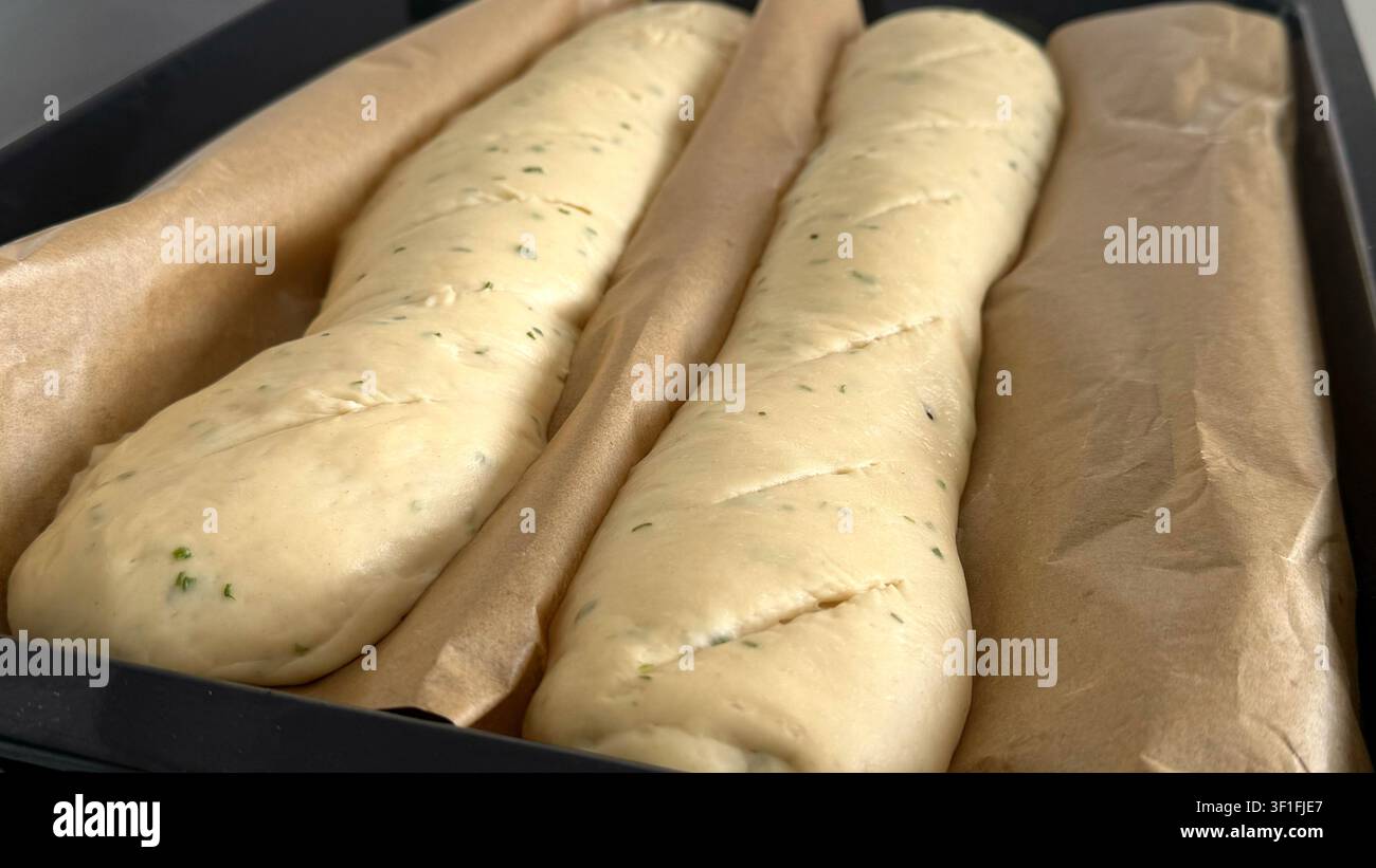 Panini di pasta appena preparati appoggiati su carta da forno in una teglia nera, pronti per il forno, con una consistenza liscia e erbe visibili mescolate dentro Foto Stock