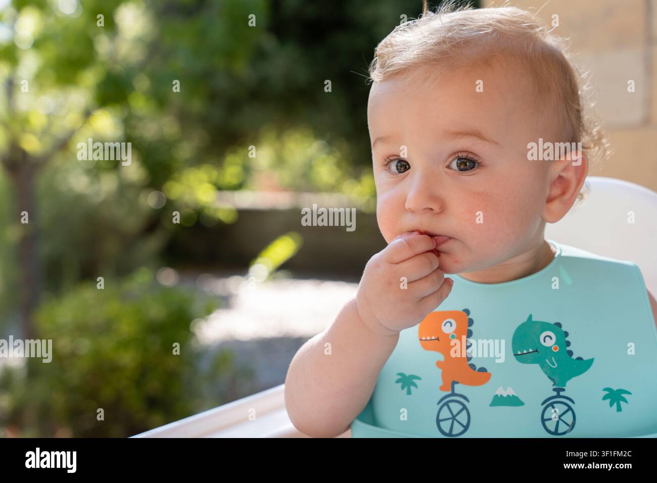 Bambino seduto all'aperto in un seggiolone, indossa un bavaglino a tema dinosauri, si nutre da solo con finger food ed esplora nuovi sapori osservando il sole verde Foto Stock