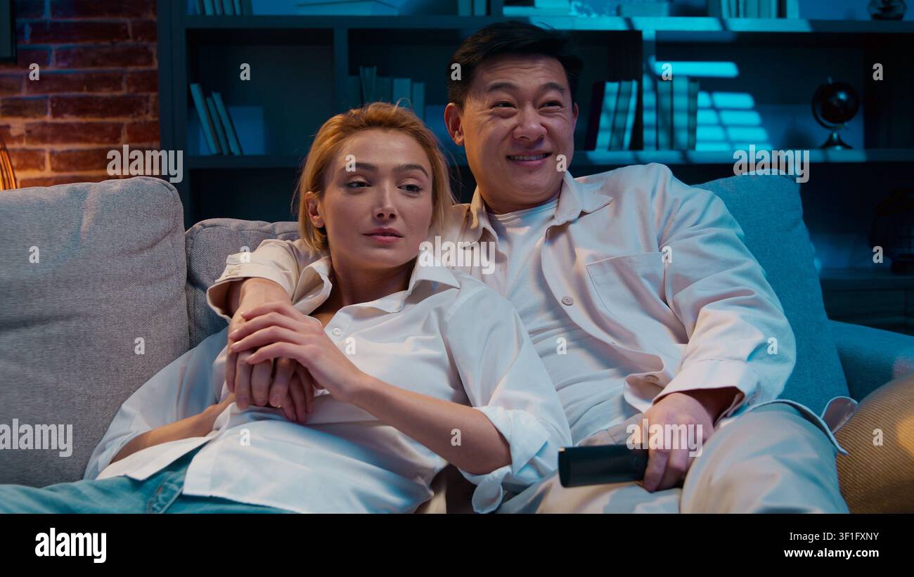 Tranquilla coppia multirazziale che guarda il cinema e la televisione coccolarsi riposando in un comodo soggiorno sul divano la sera a casa di un uomo asiatico eterogeneo Foto Stock