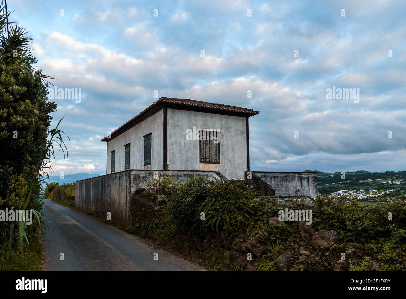 Una casa bianca con un tetto di piastrelle rosse si trova dietro un muro di cemento a Sao Miguel, nelle Azzorre, sotto un cielo nuvoloso. Foto Stock