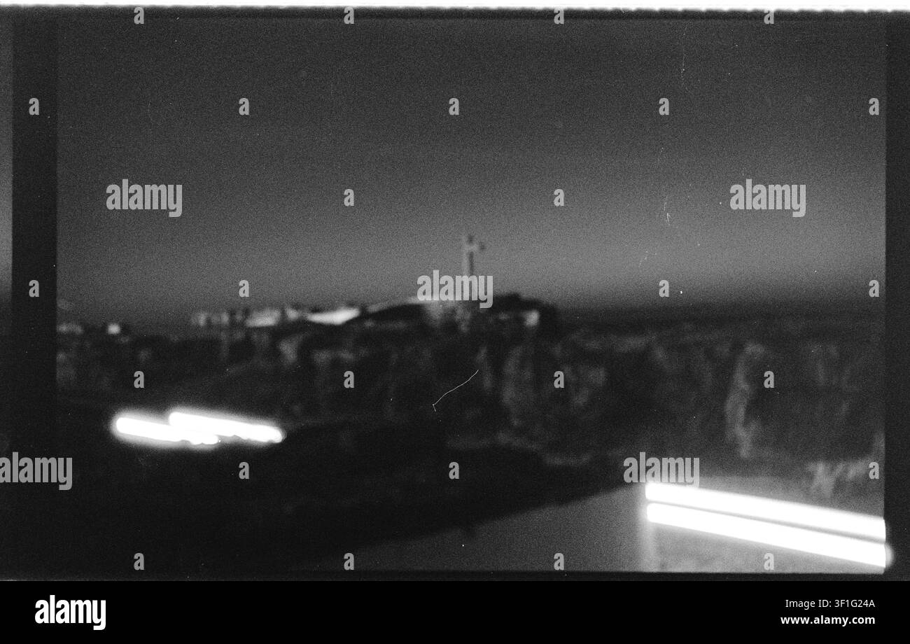 Fotografia sperimentale su pellicola in bianco e nero di un insediamento costiero sfocato con scie di luce luminose, architettura lontana e forte grana analogica Foto Stock