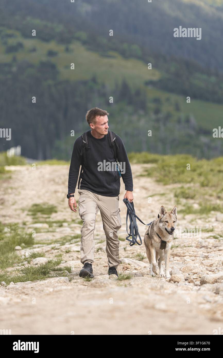 Giovane uomo che fa escursioni con il suo Wolfdog ceco su un sentiero di montagna, viaggi attivi con animali domestici e stile di vita da compagno all'aperto. Foto di alta qualità Foto Stock
