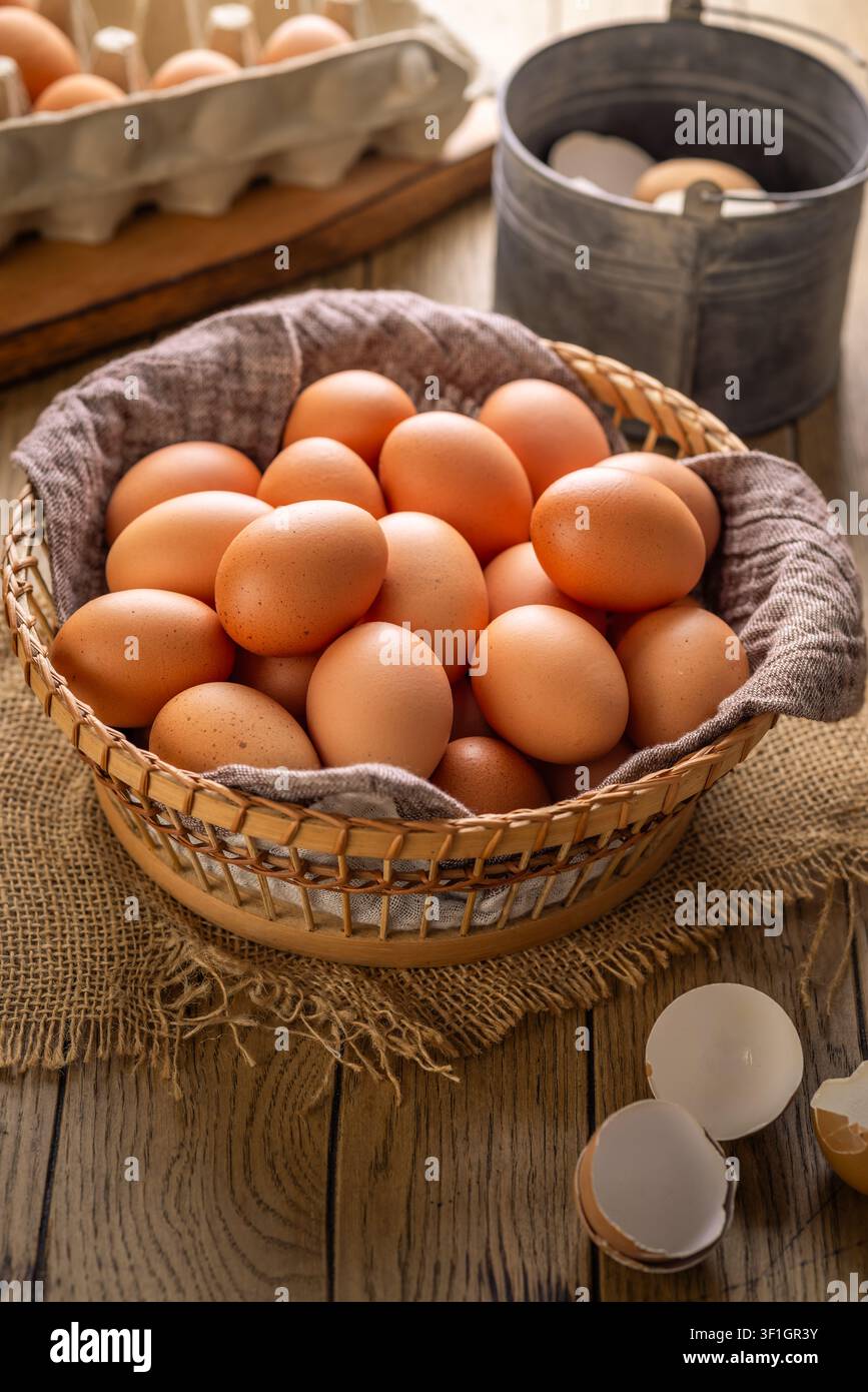 Cesto pieno di uova marroni fresche su un tavolo di legno rustico con una frusta e frammenti di guscio Foto Stock