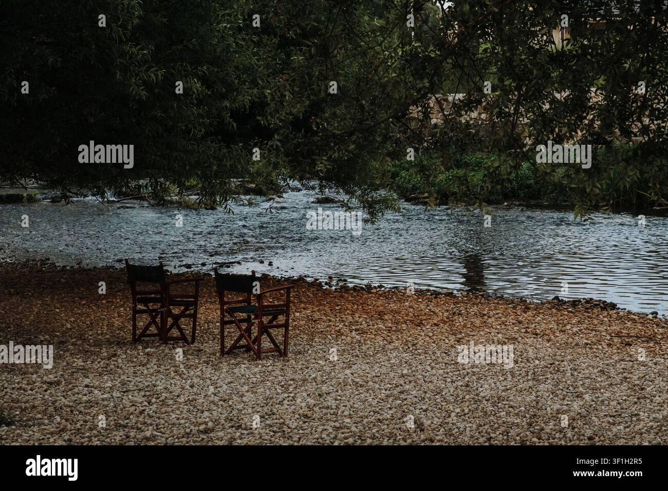 Due sedie di legno poggiano su una riva ghiaiosa sotto alberi a foglia. L'ambiente tranquillo del fiume invita a momenti di pace nella natura. Foto Stock