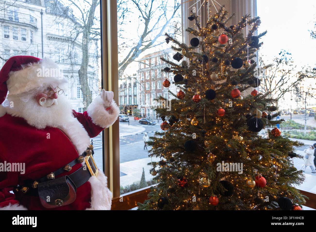 Abito di Babbo Natale con una barba bianca completa accanto ad un albero di Natale luminoso e decorato accanto ad una grande finestra che si affaccia su una strada della città. Foto Stock