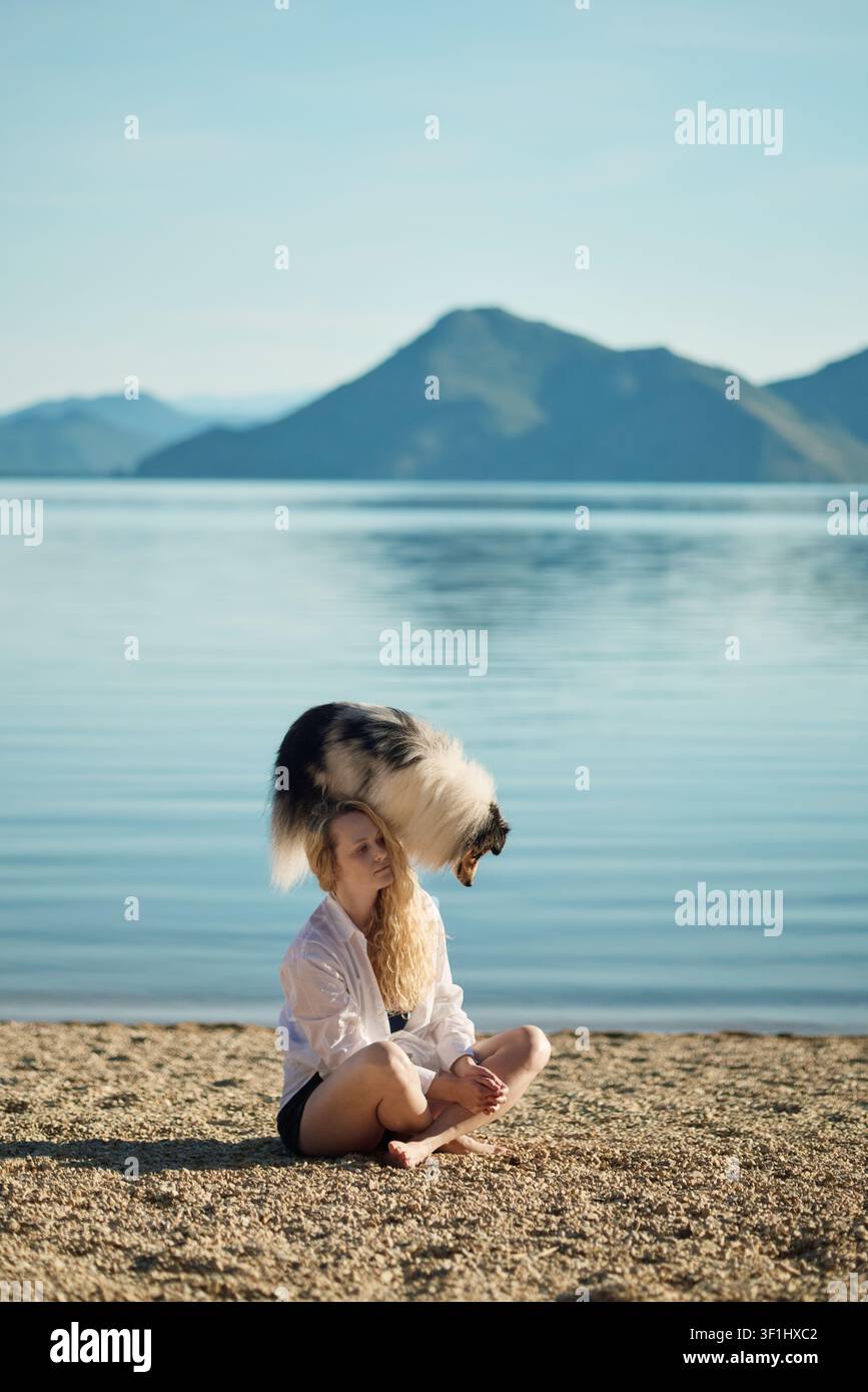 Una donna abbraccia il suo Shetland Sheepdog mentre è seduta accanto a un tranquillo lago di montagna. Bellissimo ritratto di stile di vita che esprime amore, compagnia e Foto Stock