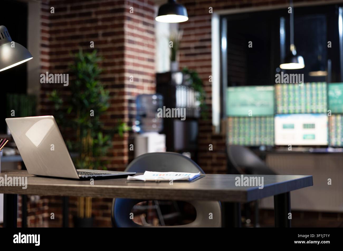Scrivania moderna vuota con un laptop e una clipboard contro un muro di mattoni di notte, con monitor sfocati che creano uno spazio di lavoro minimalista. Foto Stock
