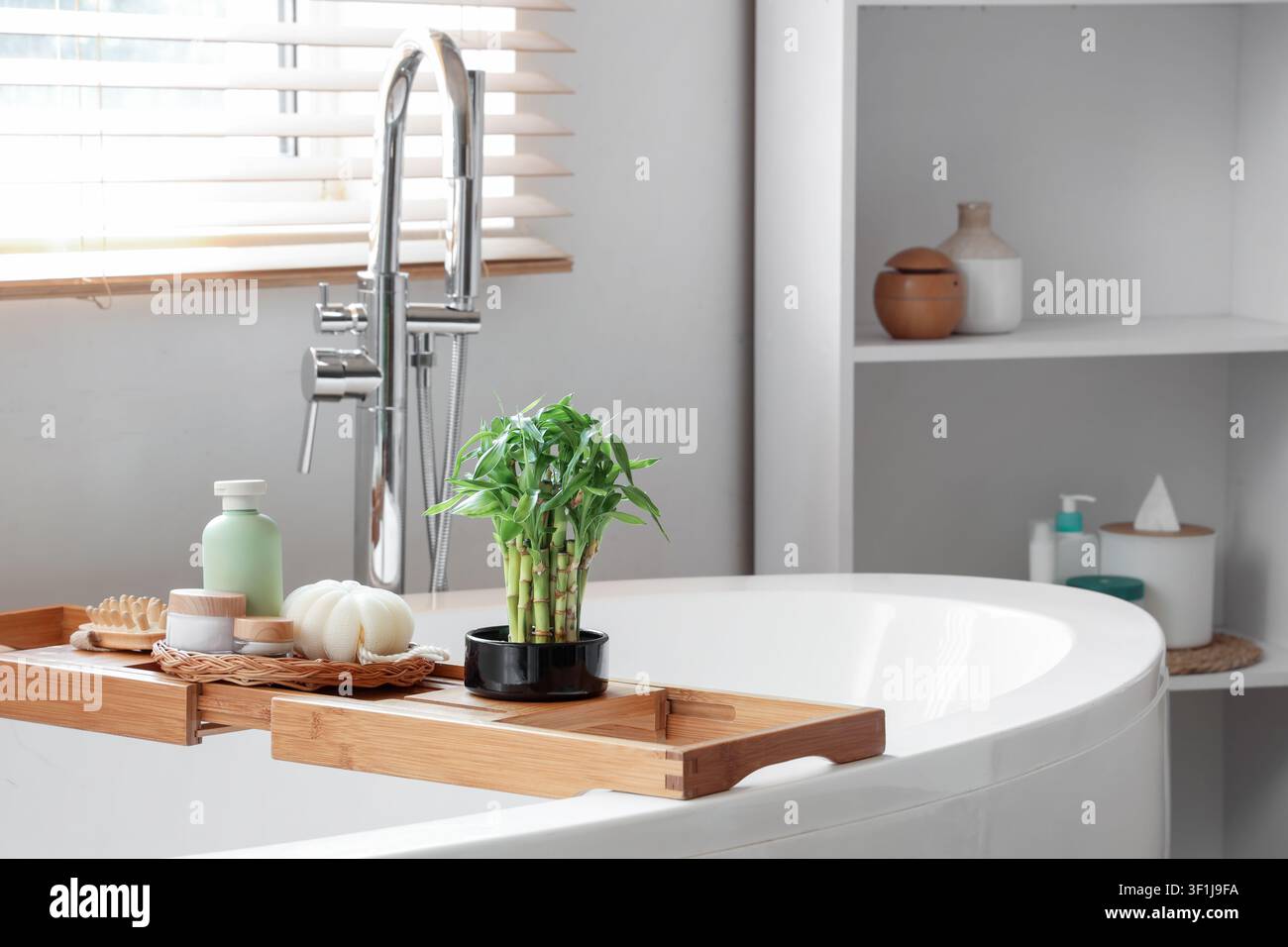 Piante di bambù e accessori per il bagno su un portapacchi di legno sopra una moderna vasca da bagno bianca in un bagno luminoso, scaffali bianchi con oggetti decorativi e articoli da toilette Foto Stock