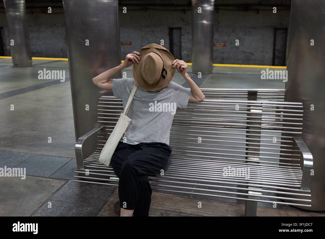 Un passeggero è seduto su una panchina nascosto dietro un cappello di paglia alla stazione della metropolitana Maverick Blue Line a East Boston, Massachusetts. Foto Stock