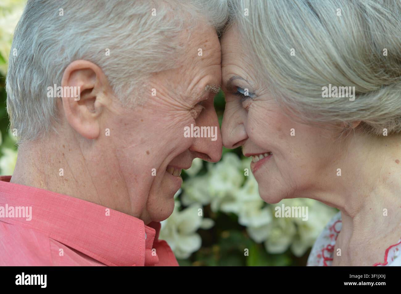 Anziano affettuoso che si strofina il naso in un lussureggiante giardino all'aperto con fiori rosa e bianchi e vegetazione, primo piano del loro momento tenero Foto Stock
