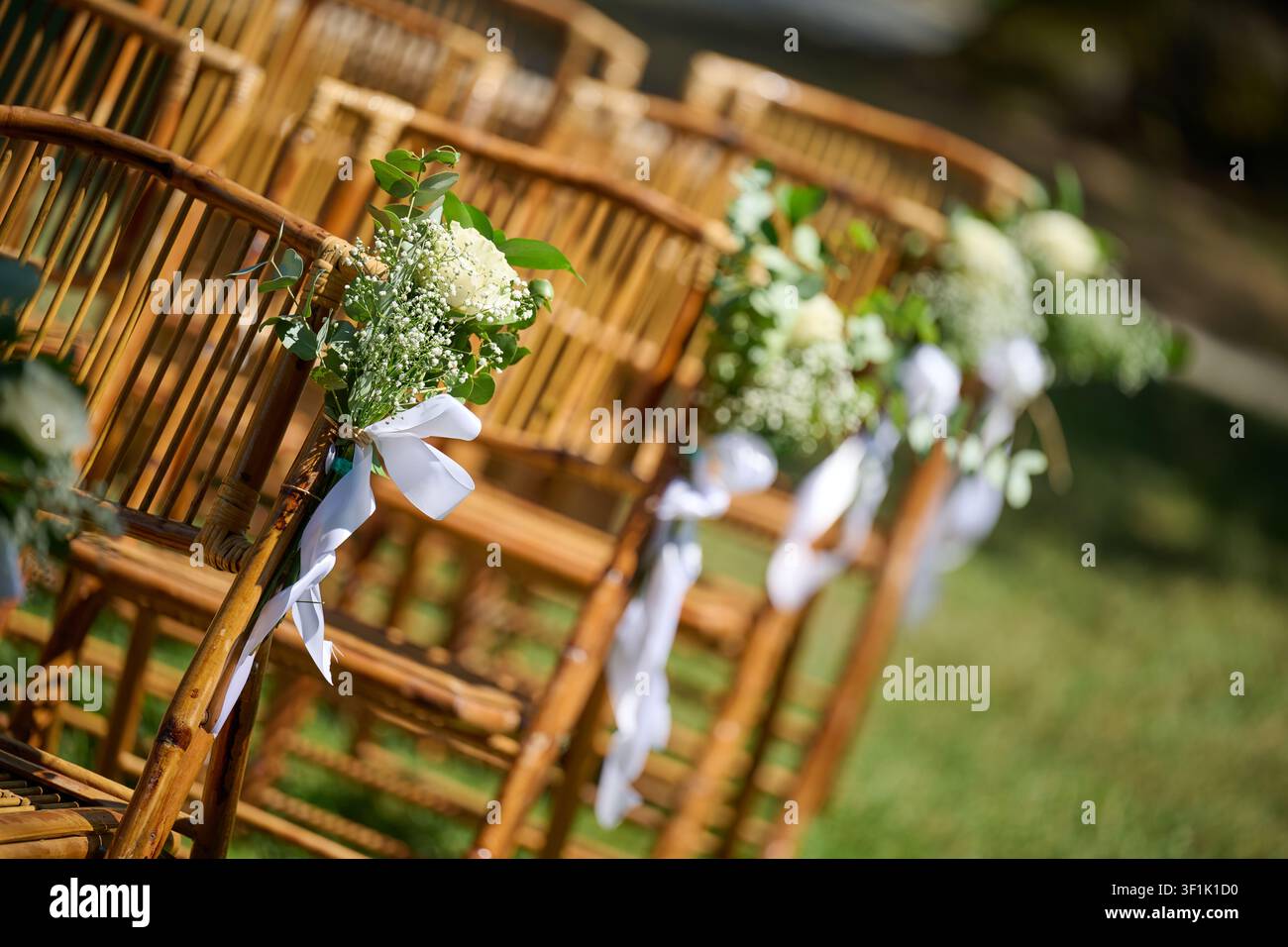 Sedie di bambù allineate sull'erba verde, ognuna adornata con un bouquet di fiori bianchi e un nastro, preparate per una cerimonia nuziale elegante all'aperto o speci Foto Stock