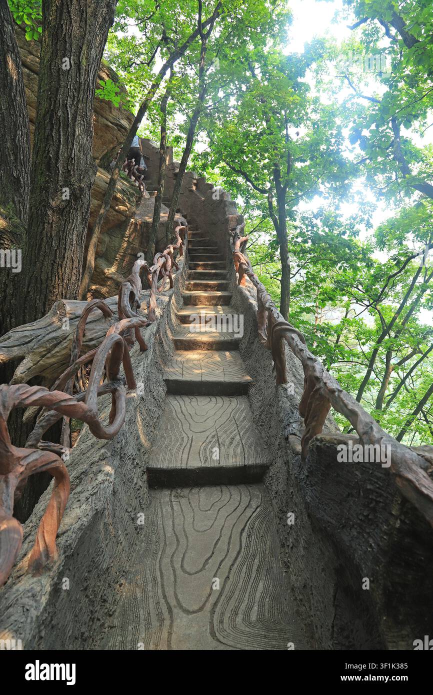 Una vista ascensionale drammatica di una scala in pietra ripida e tortuosa, progettata per imitare la texture del legno naturale, affiancata da ringhiere metalliche rustiche simili a viti. Il p Foto Stock