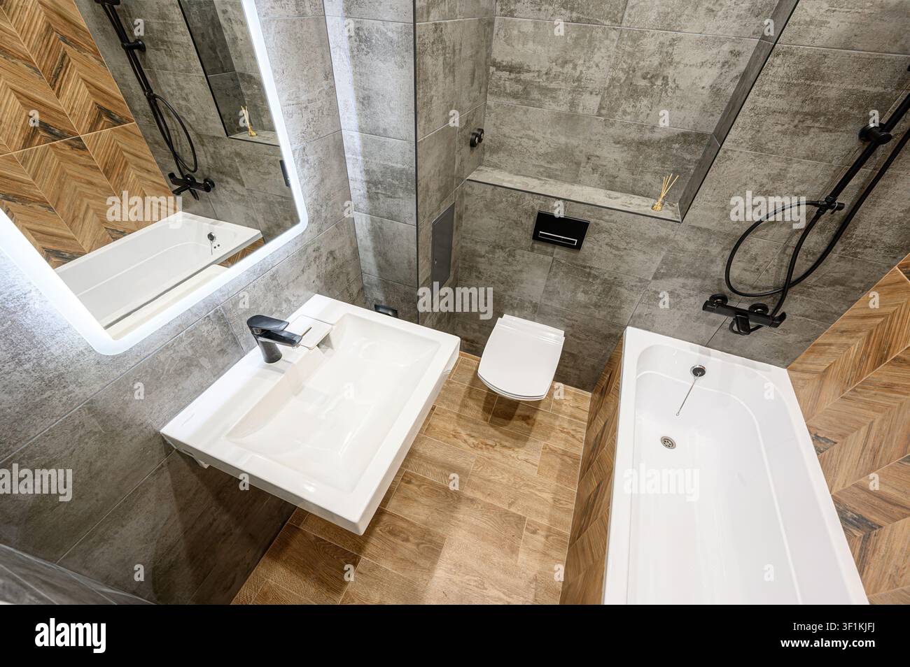 bagno moderno con pareti piastrellate grigie, un lavandino bianco, servizi igienici e vasca da bagno. Accenti di piastrelle effetto legno aggiungono calore. Gli accessori neri completano il contorno Foto Stock