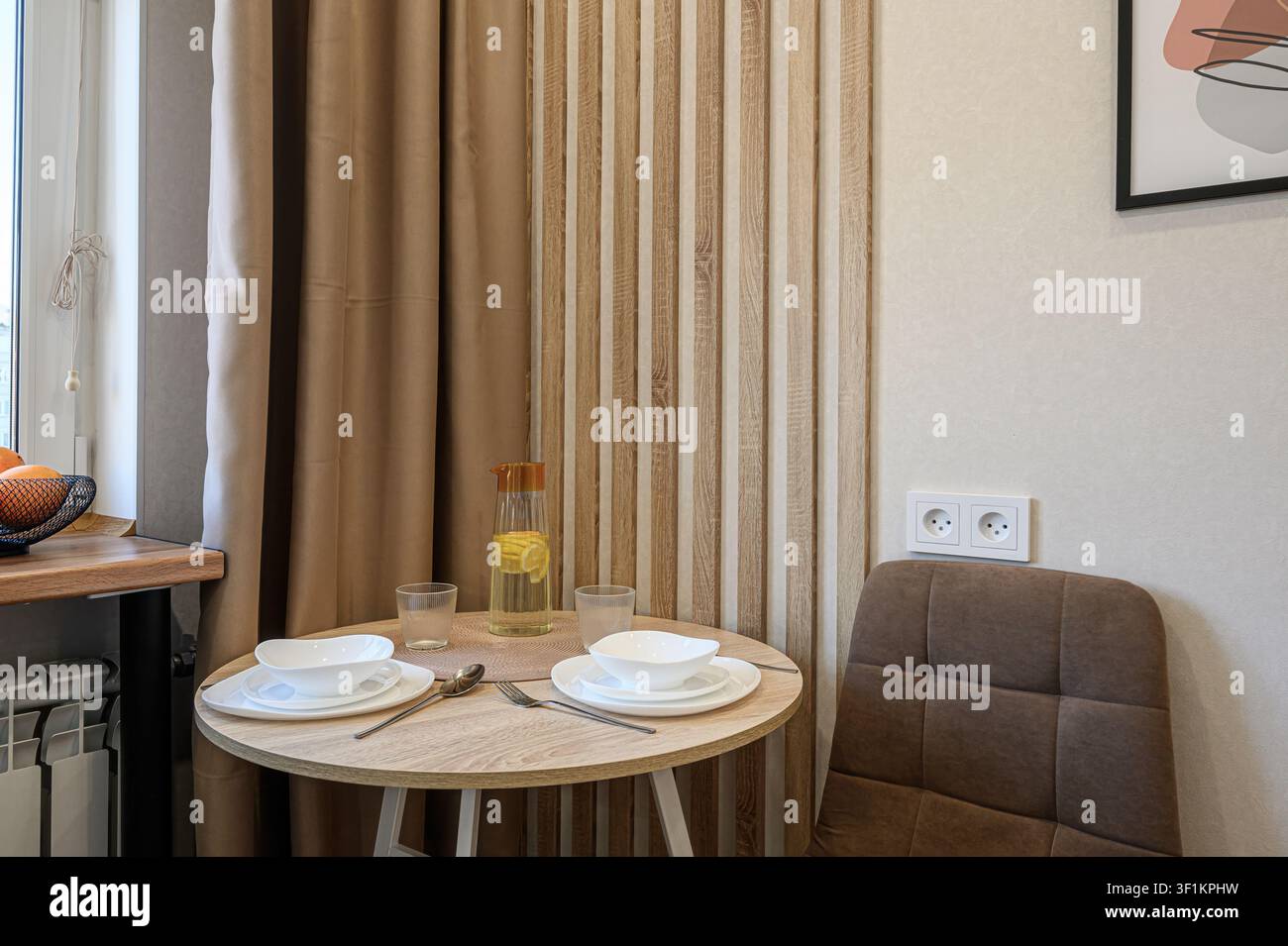 Un'accogliente zona pranzo mostra un tavolo rotondo apparecchiato per due, completo di piatti, ciotole, bicchieri e una brocca di acqua al limone, affiancato da sedie marroni. Oppure Foto Stock