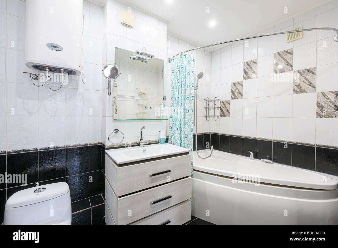 bagno luminoso con pareti piastrellate bianche e nere, un lavandino con cassetti, un WC, una vasca da bagno con tendina doccia e uno scaldabagno montato sulla parete Foto Stock