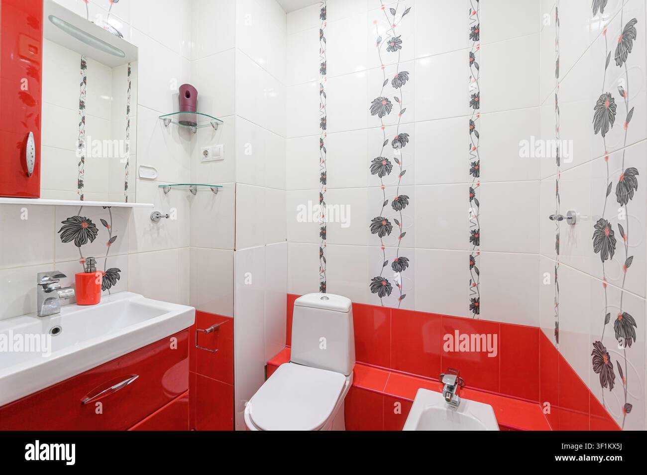 bagno luminoso con pareti piastrellate bianche con accenti floreali, un lavabo rosso, WC e bidet. Ha un design moderno e pulito Foto Stock