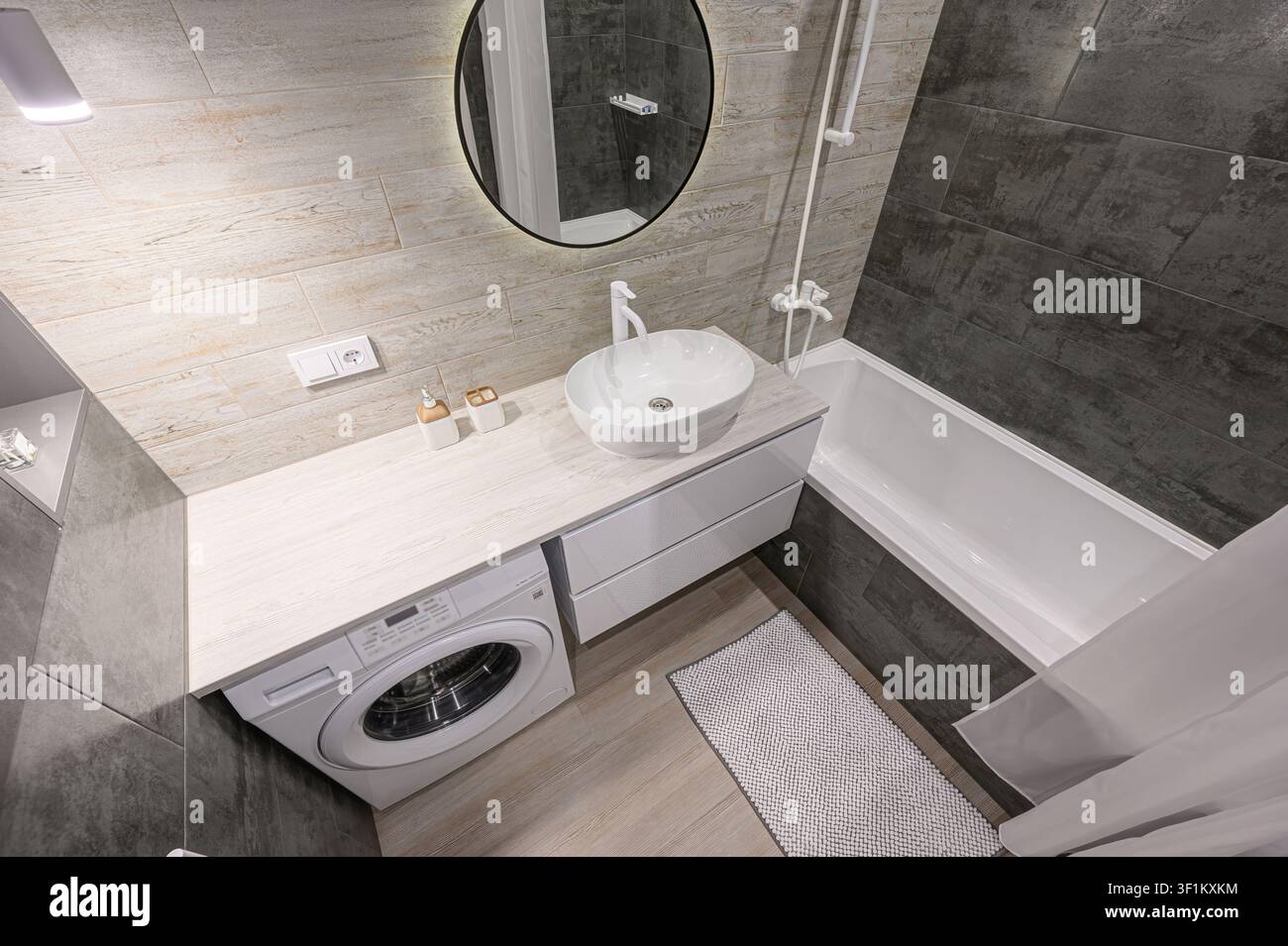 bagno moderno con piastrelle in stile legno chiaro, un lavandino bianco, una lavatrice e una vasca da bagno con piastrelle grigie Foto Stock