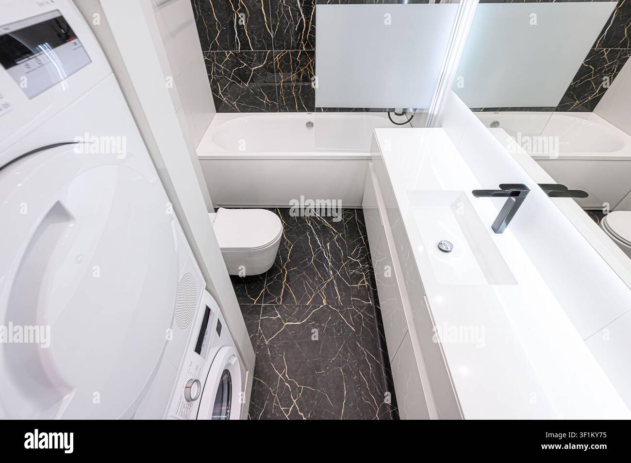 bagno moderno con sanitari bianchi, piastrelle di marmo nero e oro e un'unità lavatrice/asciugatrice impilata Foto Stock
