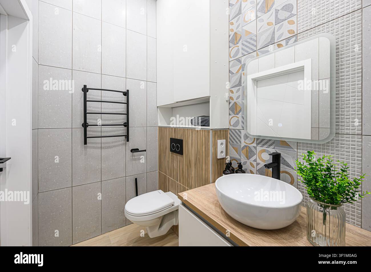 Bagno moderno con piastrelle neutre, accenti in legno, un lavandino bianco elegante e rubinetteria nera. Un muro d'accento piastrellato geometrico aggiunge un tocco stilizzato Foto Stock