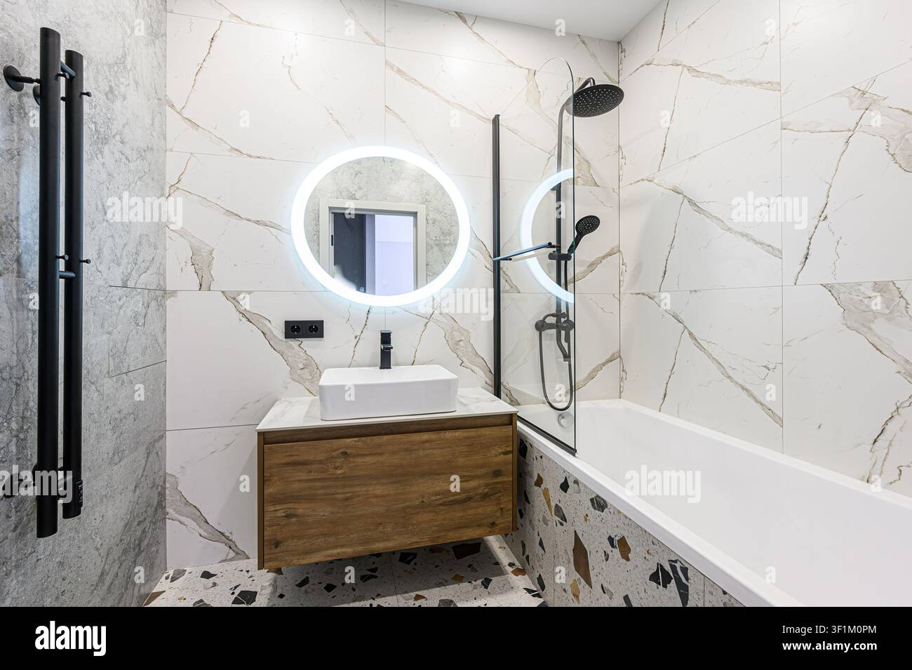 Bagno moderno con piastrelle di marmo, un lavandino sospeso in legno e uno specchio LED rotondo. I rubinetteria neri e gli accenti in terrazzo aggiungono stile Foto Stock