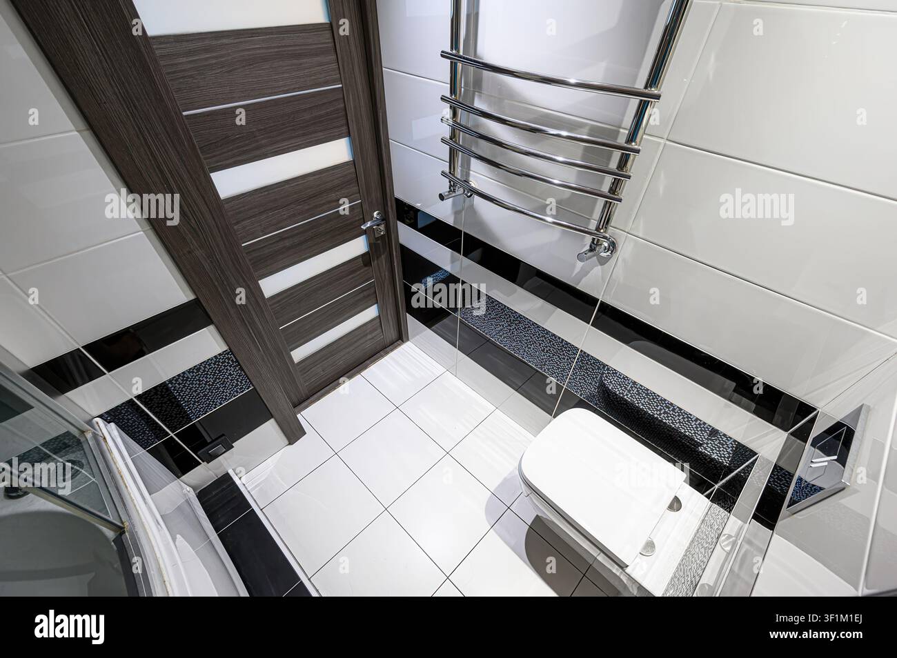 il bagno moderno presenta piastrelle bianche, una porta in legno elegante e un portasciugamani riscaldato. I dettagli neri aggiungono contrasto Foto Stock