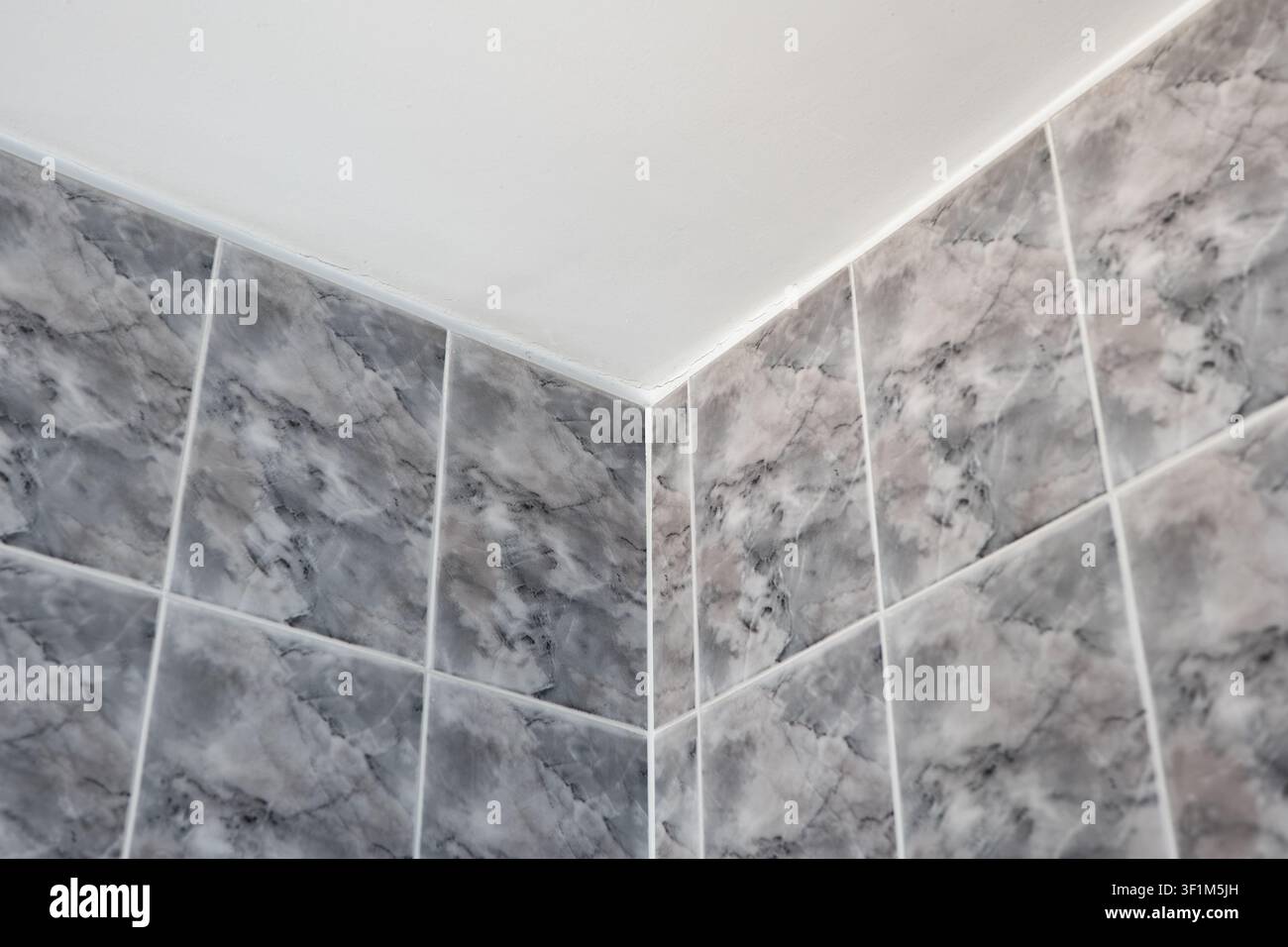 Un primo piano, scatto dall'angolo basso, che si concentra sull'angolo superiore di un bagno moderno, dove piastrelle ceramiche quadrate effetto marmo grigio incontrano una superficie pulita e semplice w Foto Stock