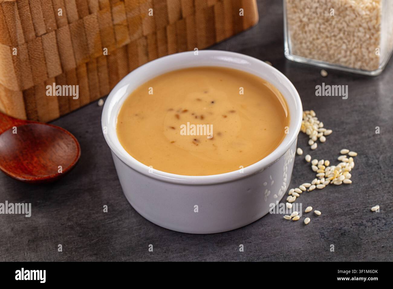 Salsa al sesamo cremosa in ciotola di ceramica bianca, circondata da semi di sesamo sparsi e cucchiaio di legno. Superficie scura e strutturata, illuminazione calda, sfondo in legno rustico Foto Stock