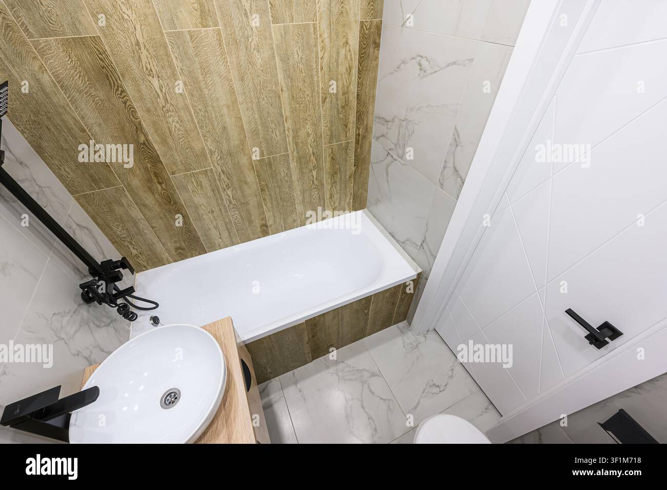 Bagno luminoso con vasca bianca, piastrelle effetto legno, dettagli in marmo e moderno rubinetto nero per la doccia. Design pulito ed elegante Foto Stock