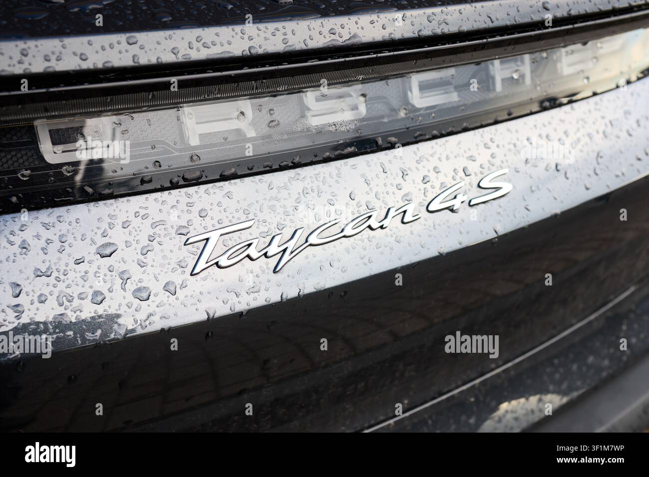 Berlino, Germania - 10 luglio 2026: Primo piano dell'emblema cromato e della barra luminosa della Porsche Taycan 4S su una carrozzeria metallica scura e bagnata con gocce di pioggia, sc Foto Stock