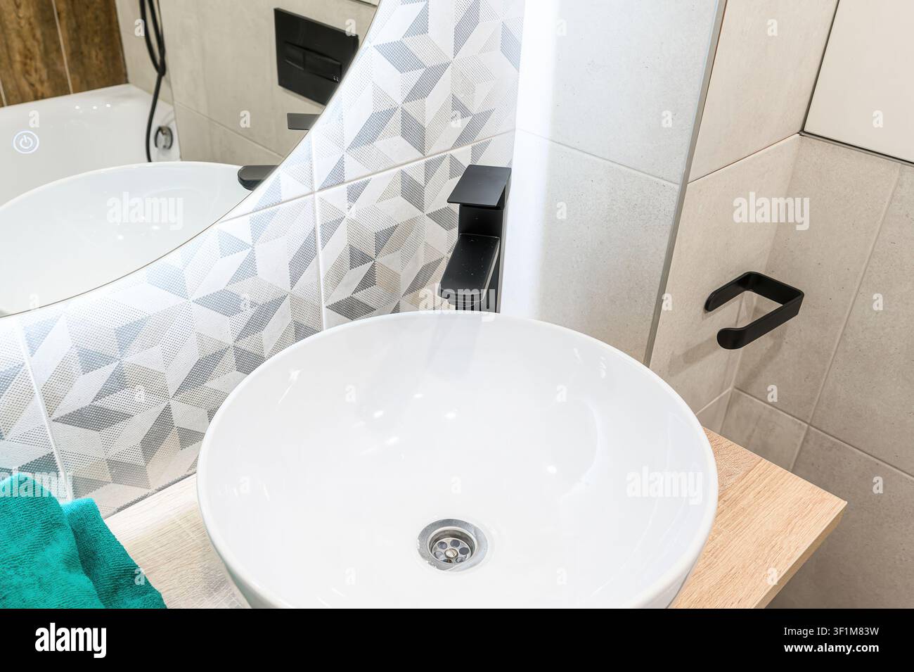 lavandino moderno da bagno con un bacino bianco e rotondo, un rubinetto nero e piastrelle a motivi geometrici. Un asciugamano blu verde è appoggiato sul piano di legno. Specchio è anche Foto Stock