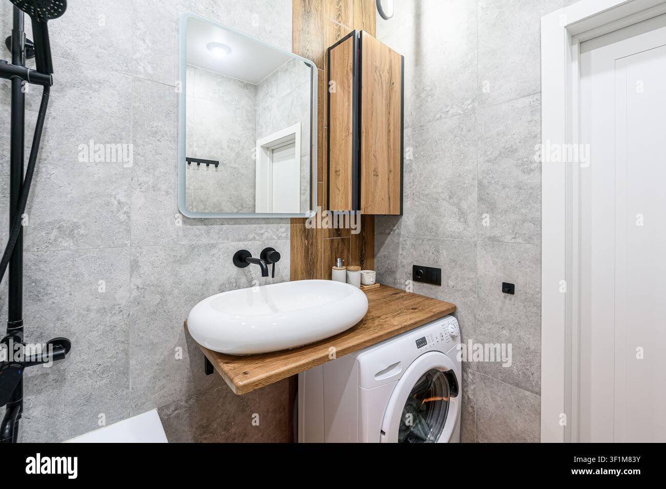 Bagno moderno con lavandino bianco, piano di lavoro in legno, lavatrice, piastrelle grigie e rubinetteria nera. Design pulito con accenti naturali Foto Stock