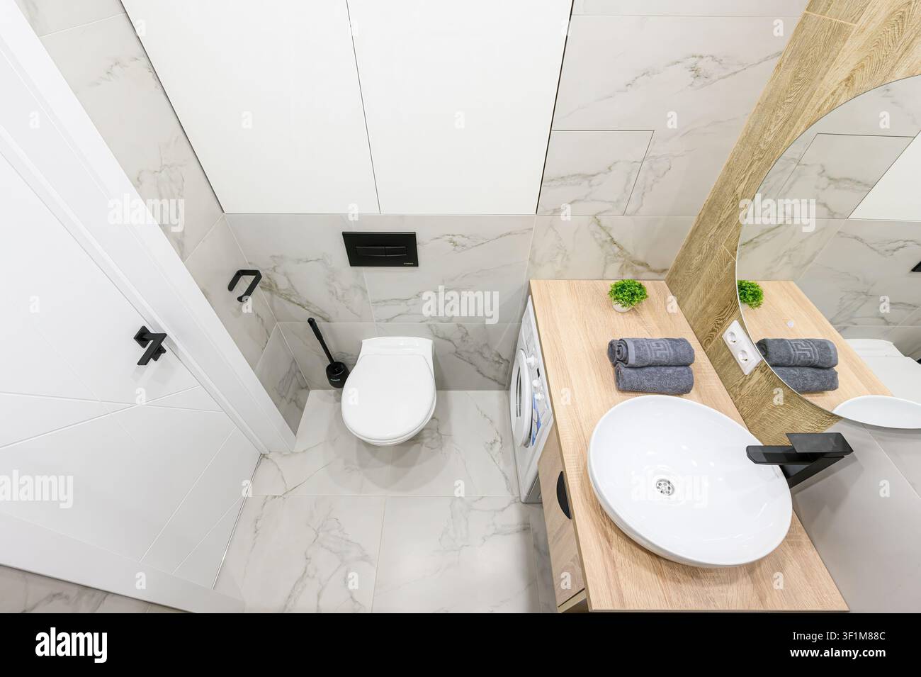 Bagno moderno con piastrelle di marmo bianco, piano di lavoro in legno, specchio rotondo, lavandino bianco, toilette e lavatrice. Accenti neri completano il design pulito Foto Stock