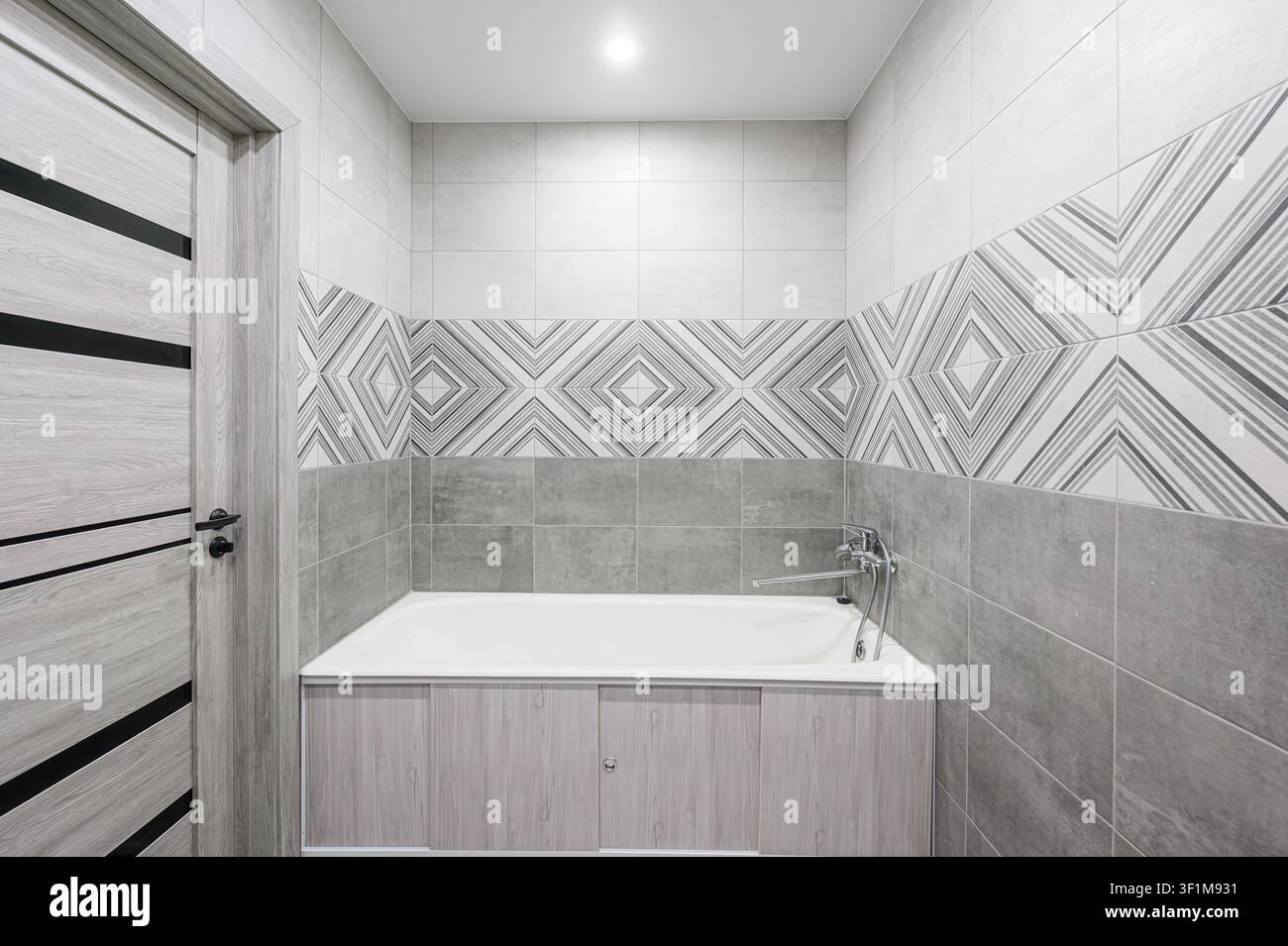 Bagno moderno con piastrelle grigie, un accento a motivi, una vasca bianca e una porta a venatura leggera con dettagli neri Foto Stock