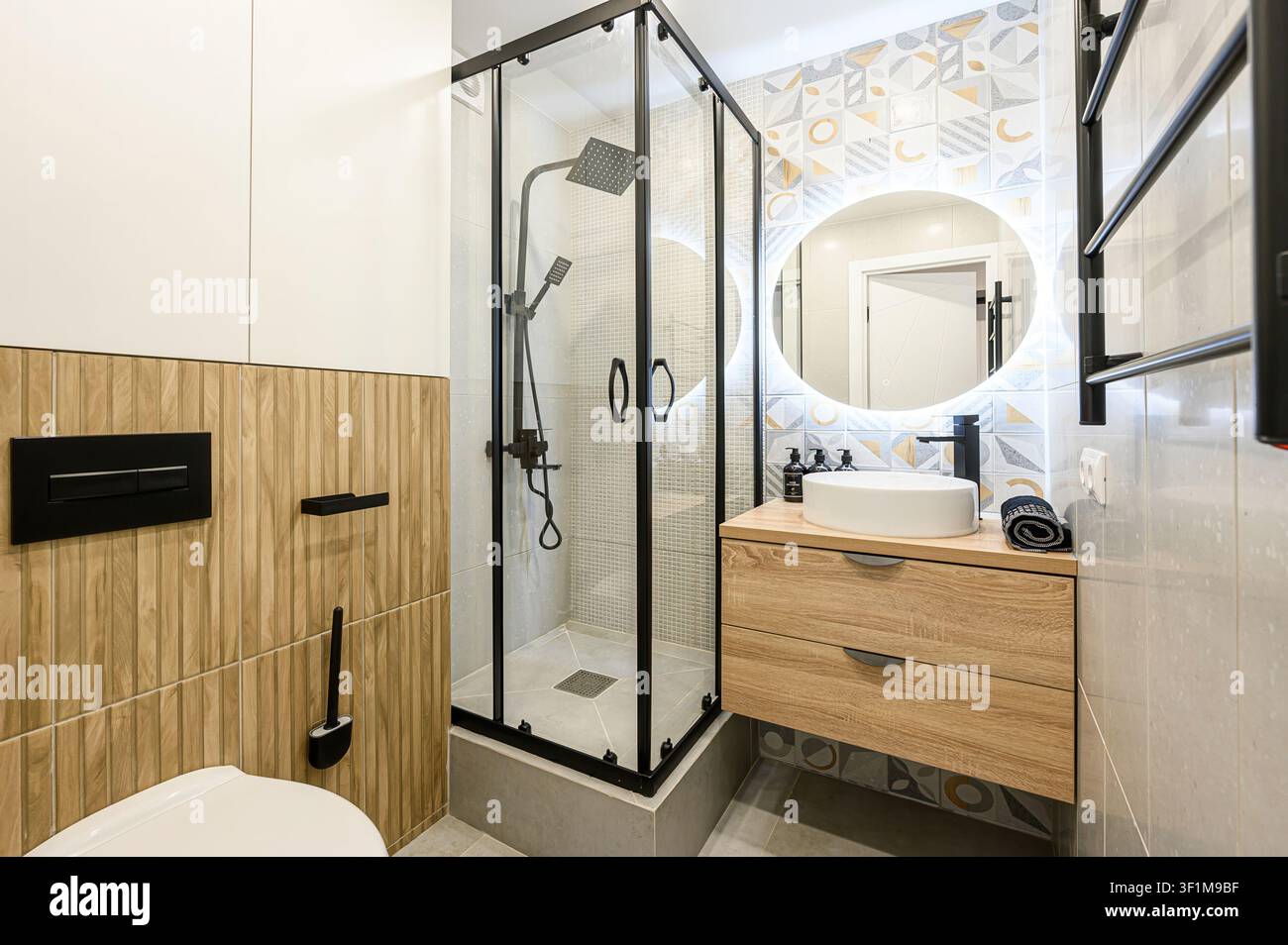 Bagno moderno con doccia in vetro, mobiletto in legno, specchio LED rotondo e accenti di piastrelle geometriche Foto Stock