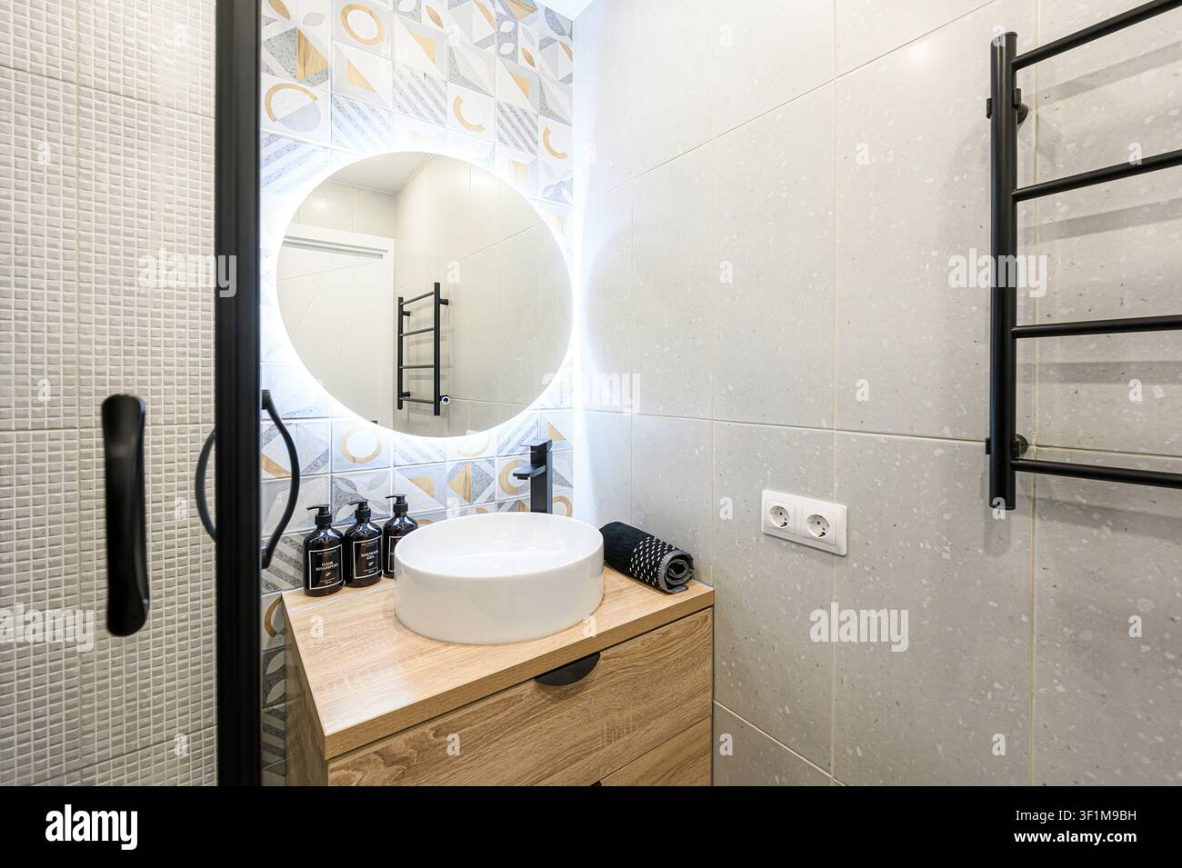 Il bagno moderno presenta uno specchio rotondo retroilluminato sopra un lavandino bianco. Accenti neri contrastano con mobili in legno chiaro e piastrelle decorate Foto Stock