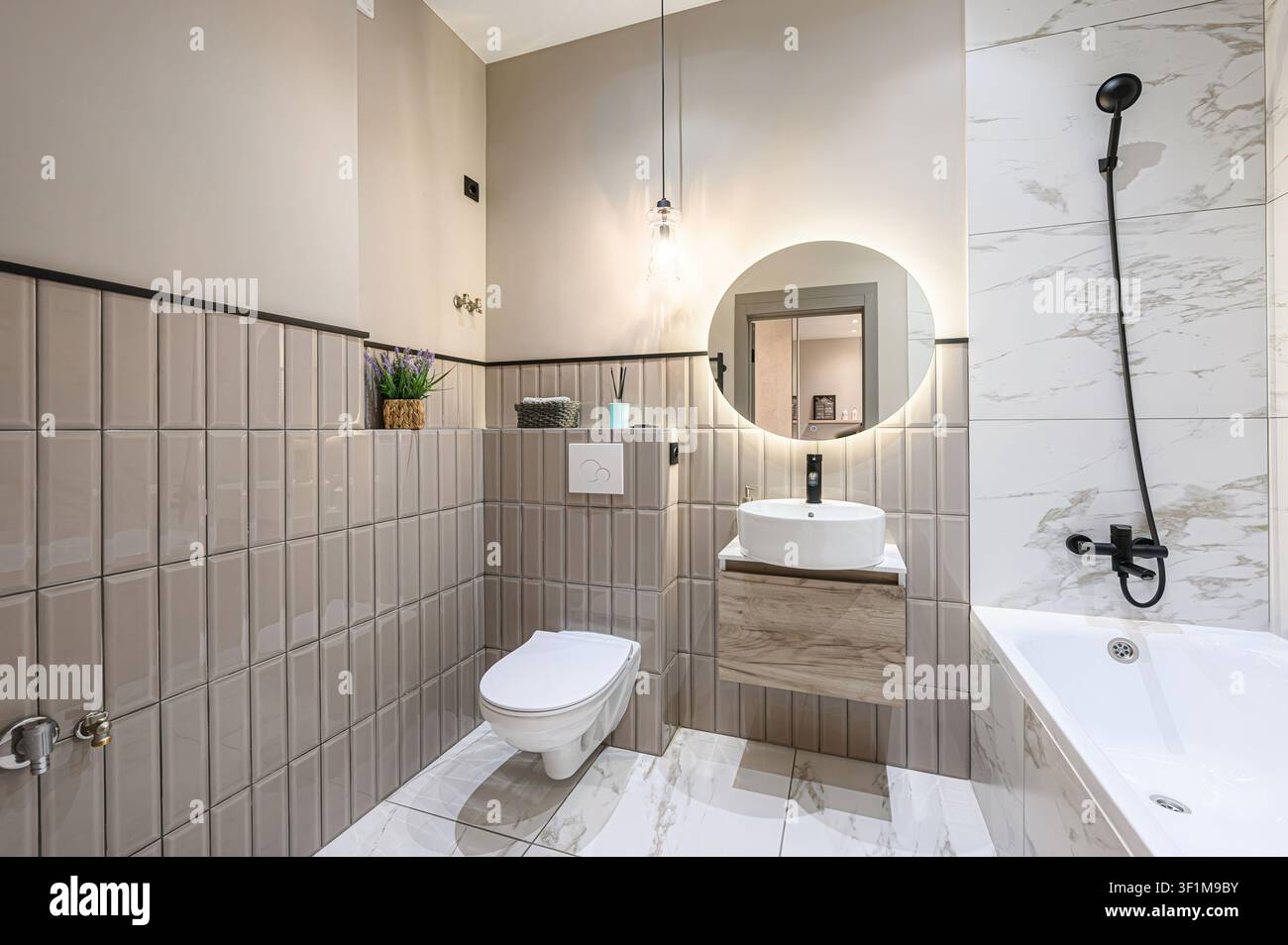 Bagno moderno con piastrelle grigie, lavandino bianco, specchio LED rotondo e vasca bianca con rubinetteria nera. Design pulito e contemporaneo Foto Stock
