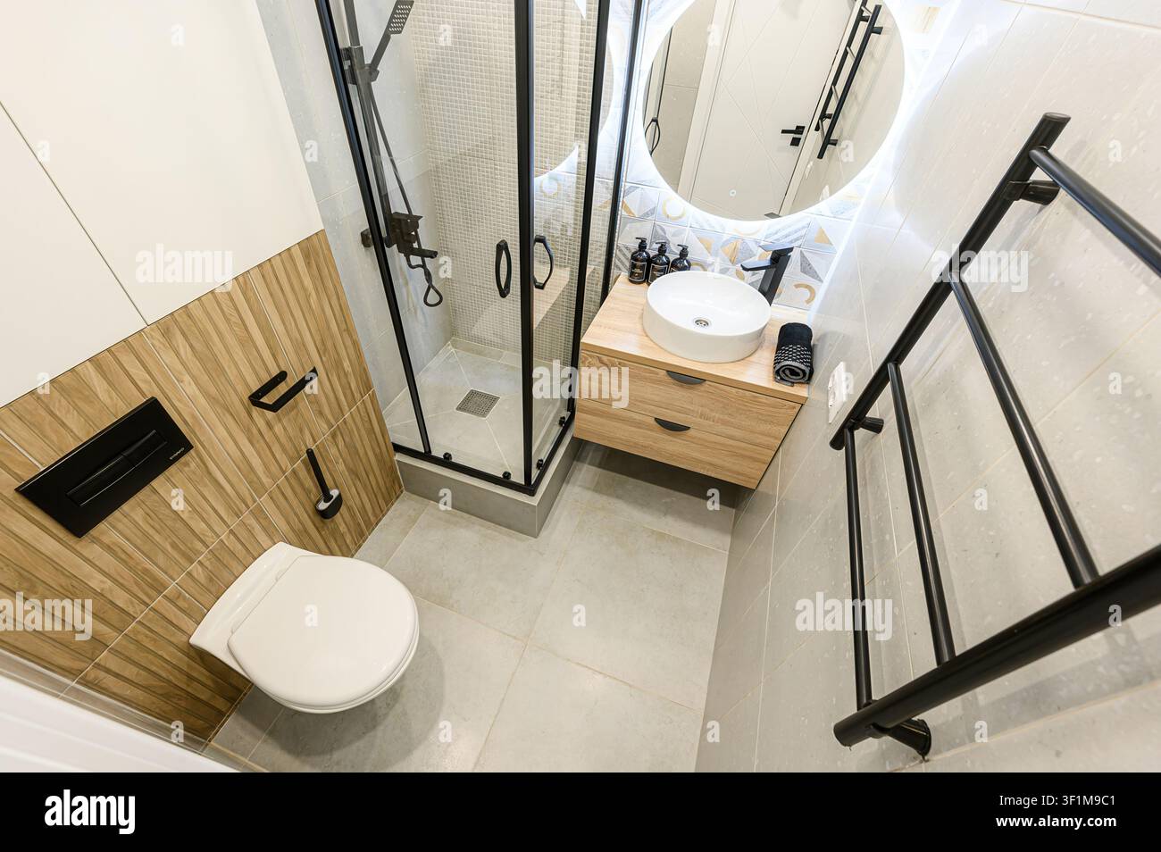 Bagno moderno con doccia in vetro, mobiletto in legno, specchio LED rotondo e accenti di piastrelle geometriche Foto Stock