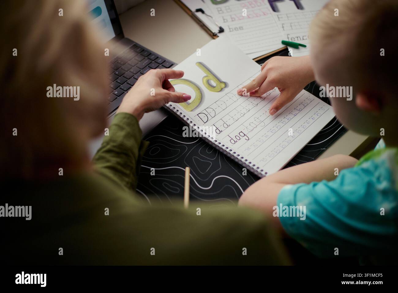 Bambino che impara la calligrafia con la guida di un adulto, tracciando la lettera d in un quaderno accanto a un laptop e a schede fonetiche. Utile per l'educazione alla alfabetizzazione, homeschooling, promozione del tutoraggio Foto Stock