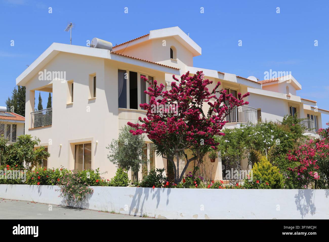 Una villa a due piani color crema con balconi è incorniciata da un bougainvillea rosa in piena fioritura e da un lussureggiante giardino a Cipro. Foto Stock