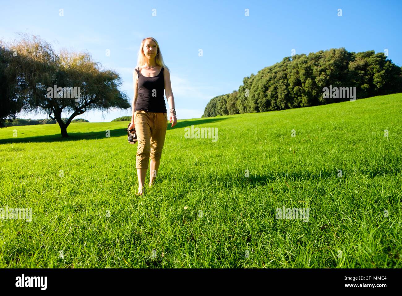 Donna che cammina a piedi nudi su un prato verde portando sandali, godendosi la natura e la libertà Foto Stock