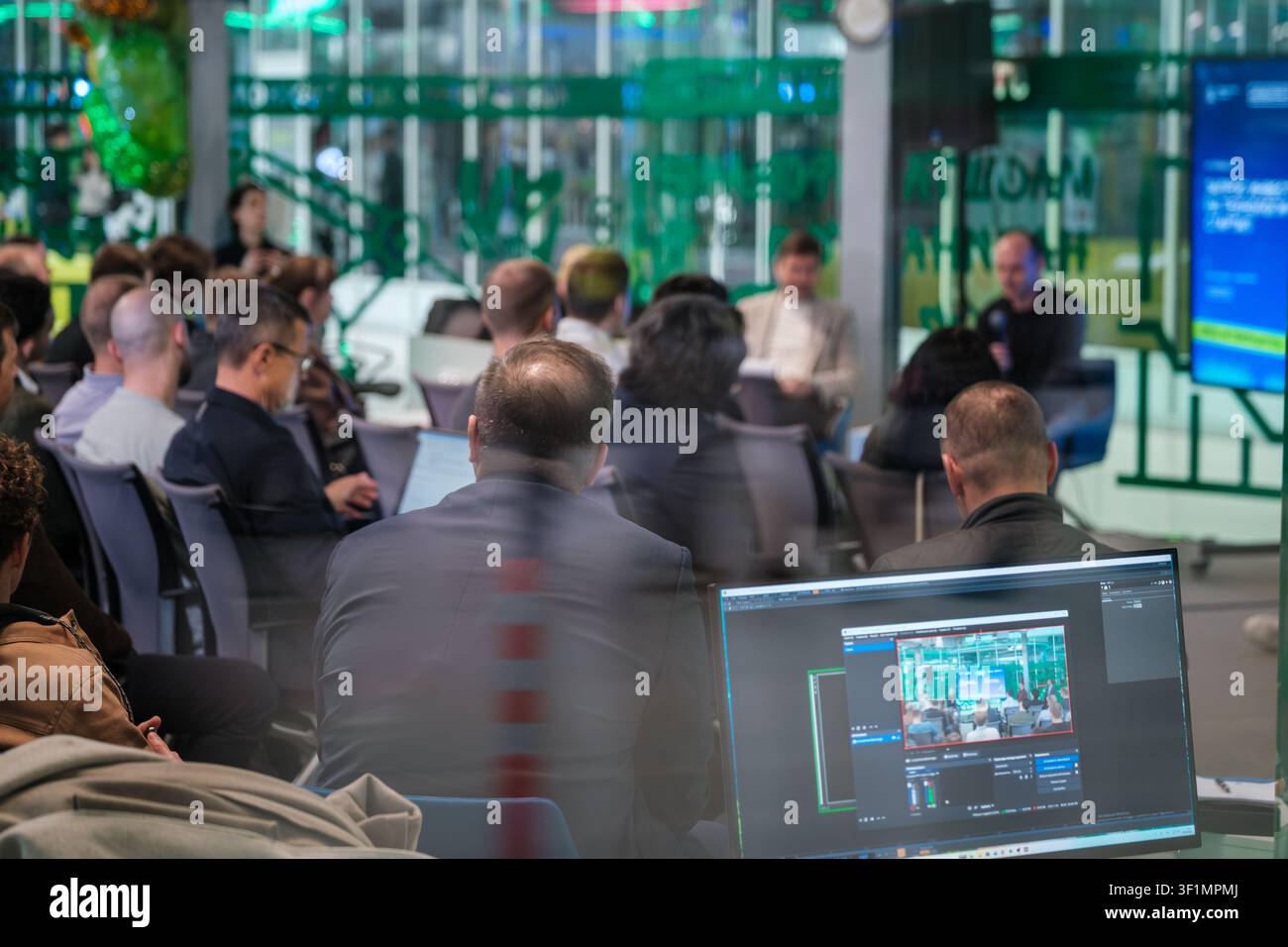 Pubblico di una conferenza aziendale che osserva una discussione di pannello in un ufficio moderno, il primo piano sfocato mostra un laptop in live streaming e partecipanti che prendono appunti. Foto Stock