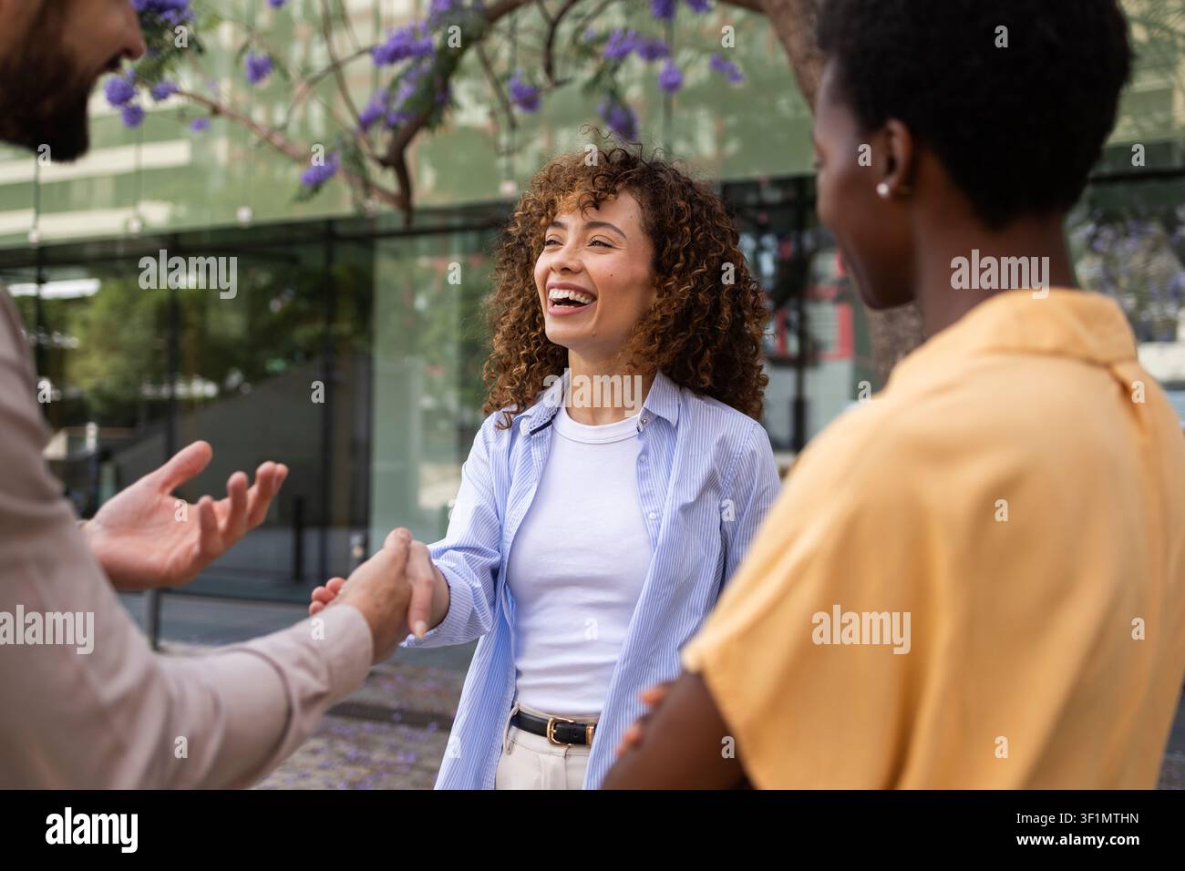 Colleghi sorridenti si salutano con una stretta di mano durante un incontro all'aperto vicino ad edifici moderni di uffici, mostrando lavoro di squadra, networking e positività Foto Stock
