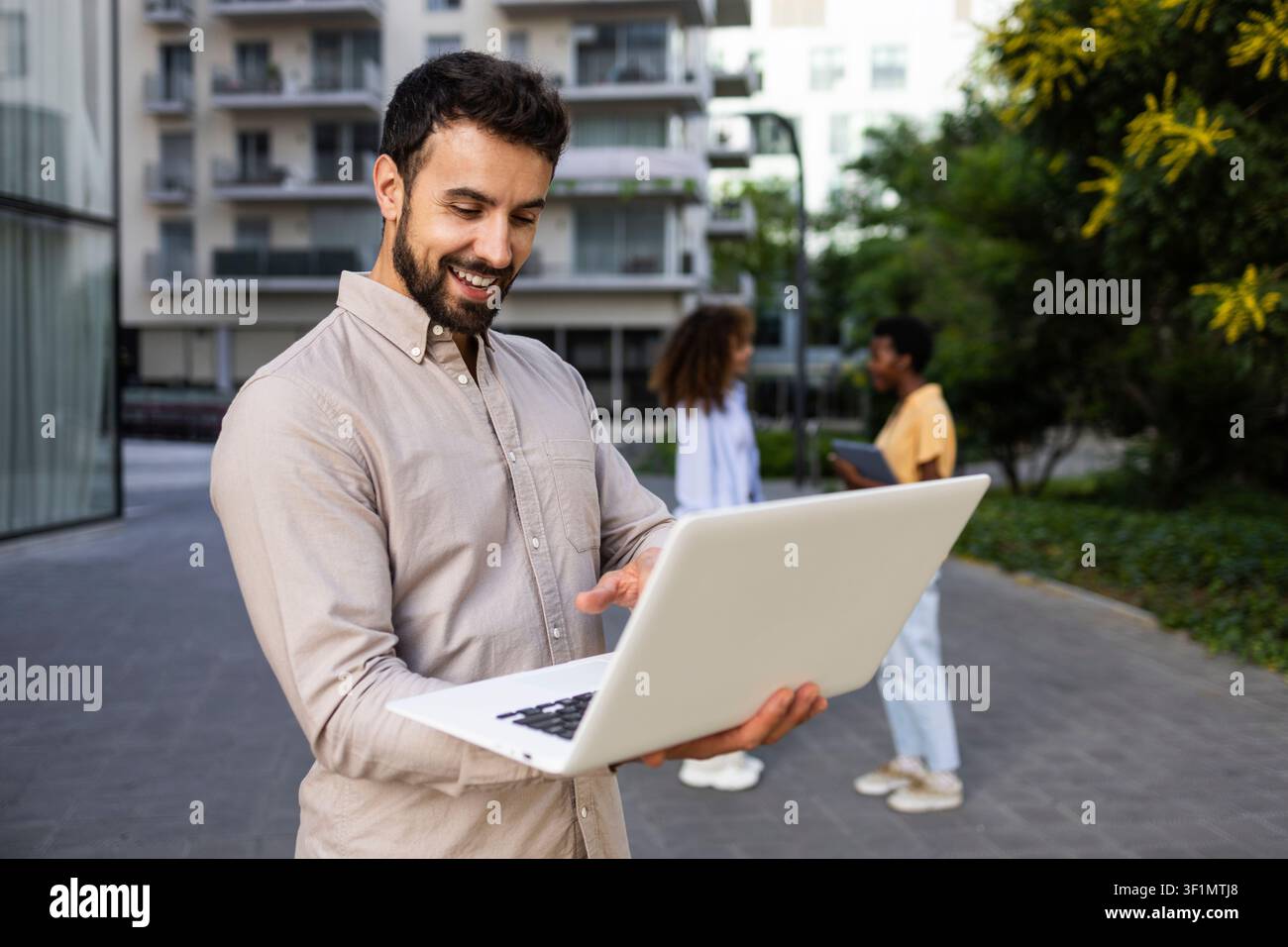 Giovane uomo sorridente che lavora su un laptop all'aperto in un complesso residenziale moderno, utilizzando l'internet senza fili in una giornata di sole mentre persone passano in primo piano Foto Stock