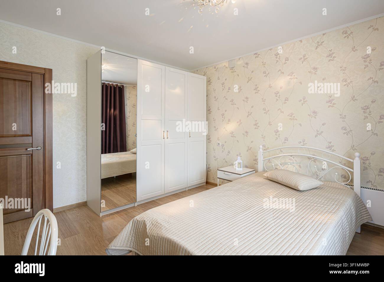 camera luminosa con carta da parati floreale presenta un letto bianco in ferro battuto, un comodino abbinato e un grande armadio bianco con una porta a specchio. Il fl Foto Stock