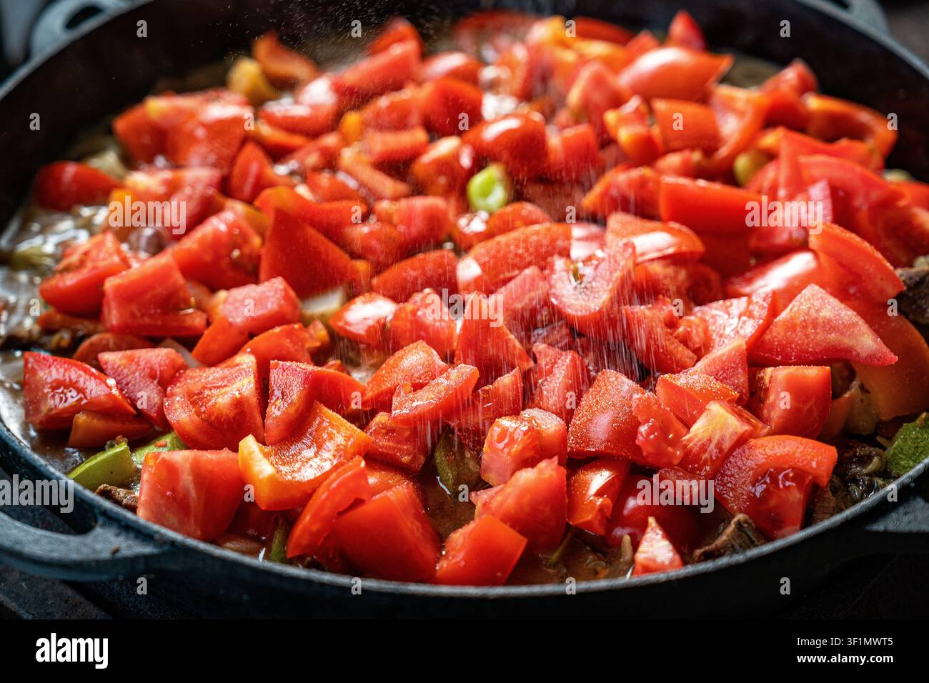 Primo piano di pomodori e peperoni a cubetti in una padella, arricchiti con una spolverata di condimento. I vivaci colori rossi e verdi evidenziano la freschezza o Foto Stock
