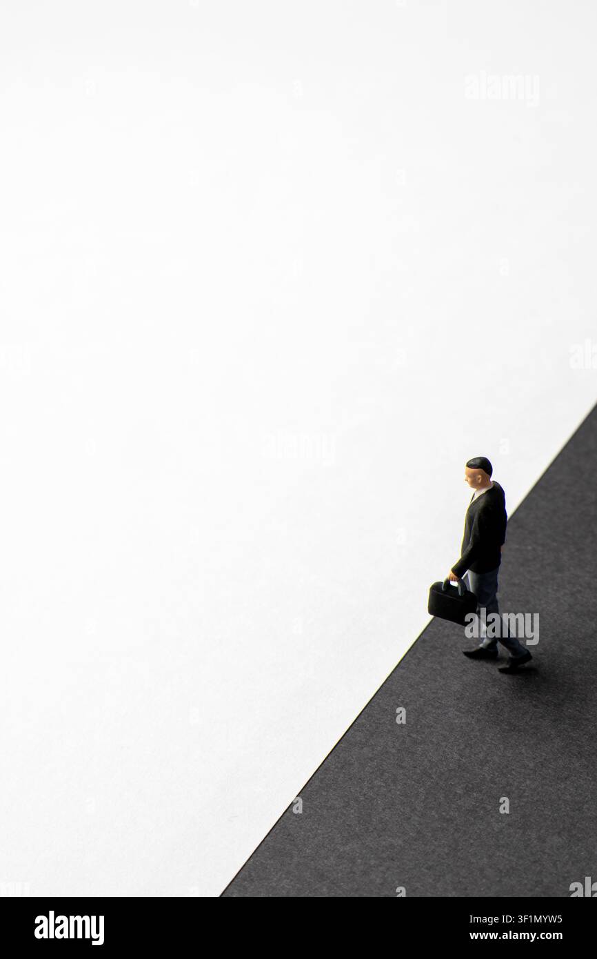 Uomo d'affari in miniatura con una valigetta che cammina lungo una linea di divisione diagonale tra sfondi neri e bianchi. Transizione, rischio Foto Stock