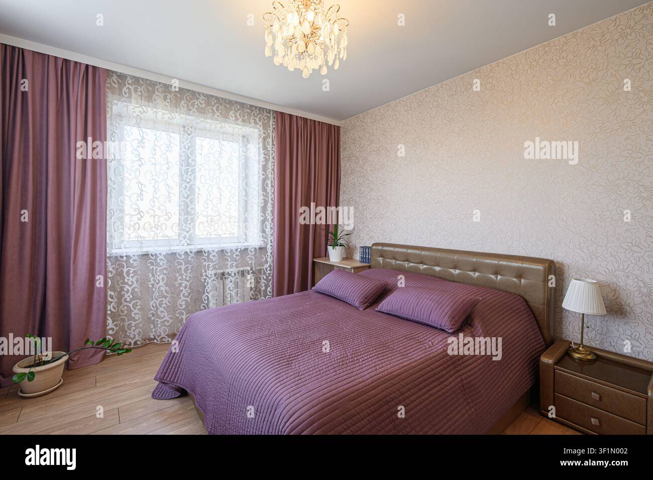 Camera da letto accogliente con letto viola, tende coordinate, carta da parati a motivi e un lampadario di cristallo per un'atmosfera calda e invitante Foto Stock